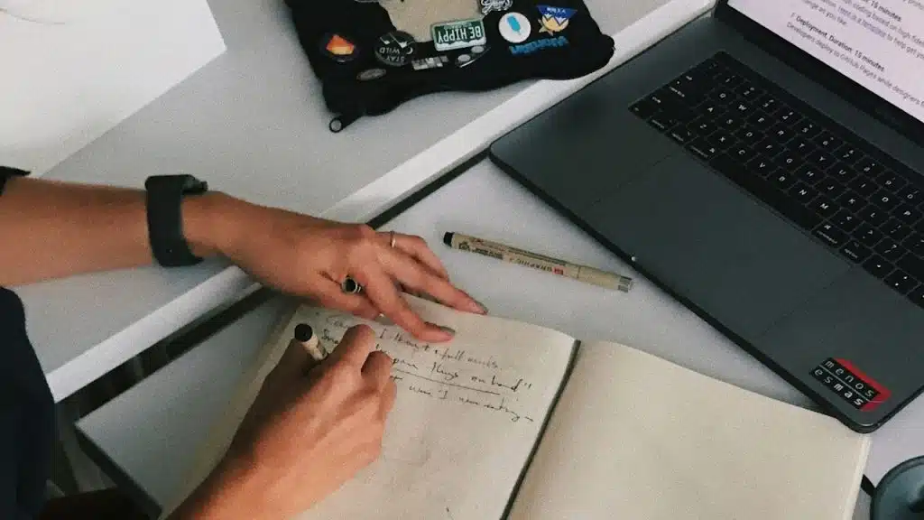 A man writing in a journal with a laptop placed near it.
