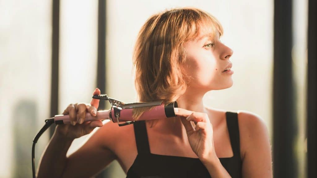 A woman curling her hair with a curling iron near a window.