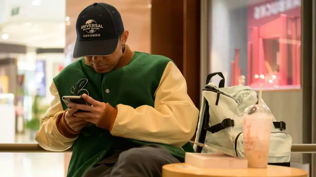 A man wearing a cap and baseball jersey using his mobile while seated at a table.
