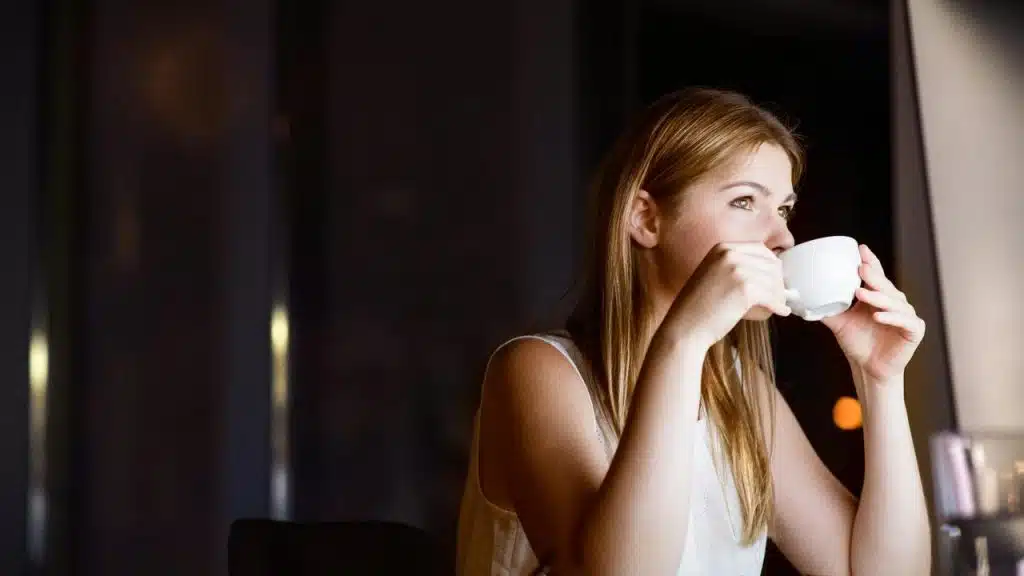 A woman sitting by a window drinking from a white cup.