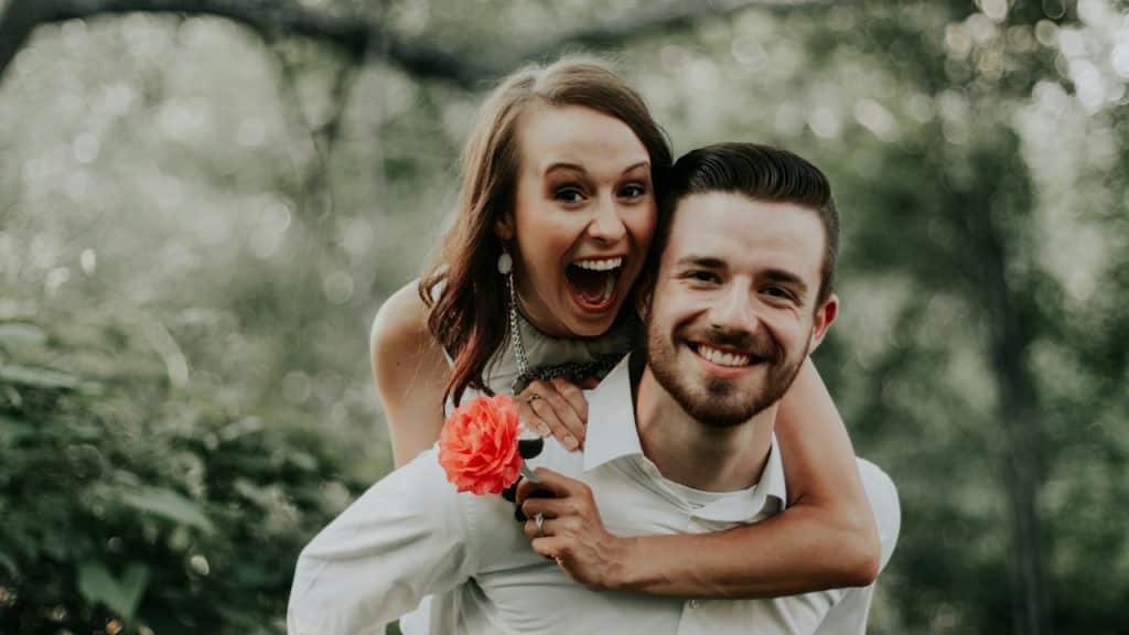 A laughing woman holding a flower being carried by a smiling man on his back.