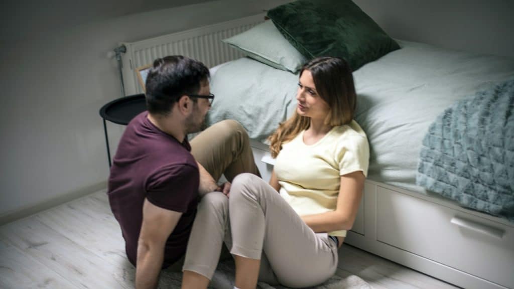 A couple sitting on the floor in a bedroom and staring at each other.