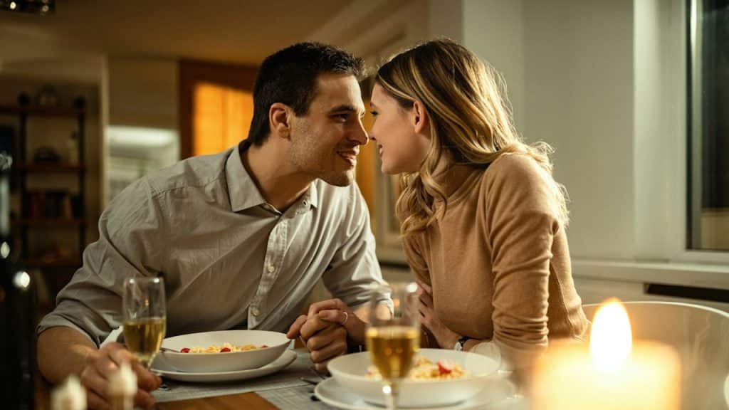 A Couple leaning in for a kiss while having dinner and wine at a table.