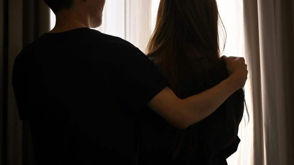 A man with his arm around a woman as they stand together by a window.