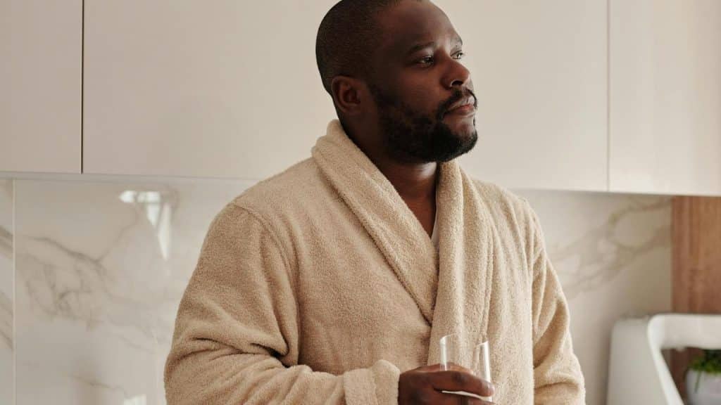 A man in a bathrobe holding a glass of water in a kitchen.