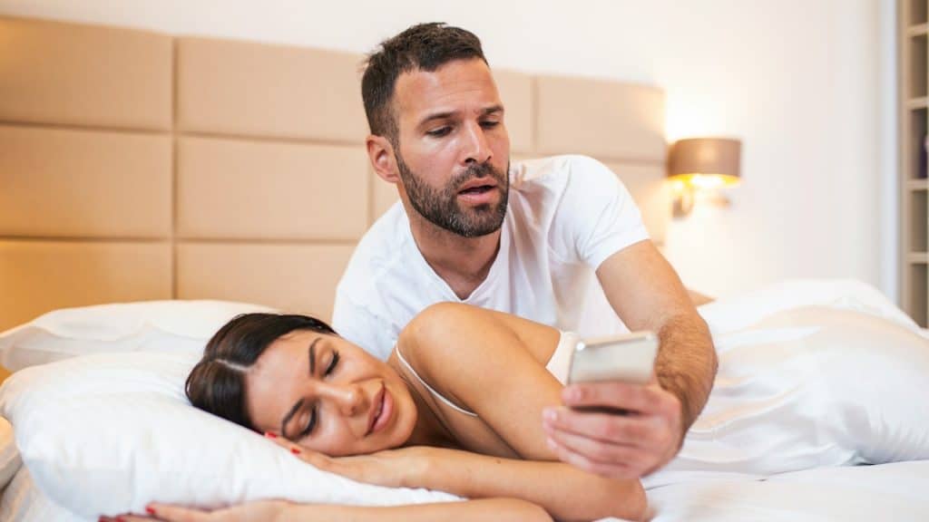 A bearded man leans over a sleeping woman in bed, looking at something on a smartphone.