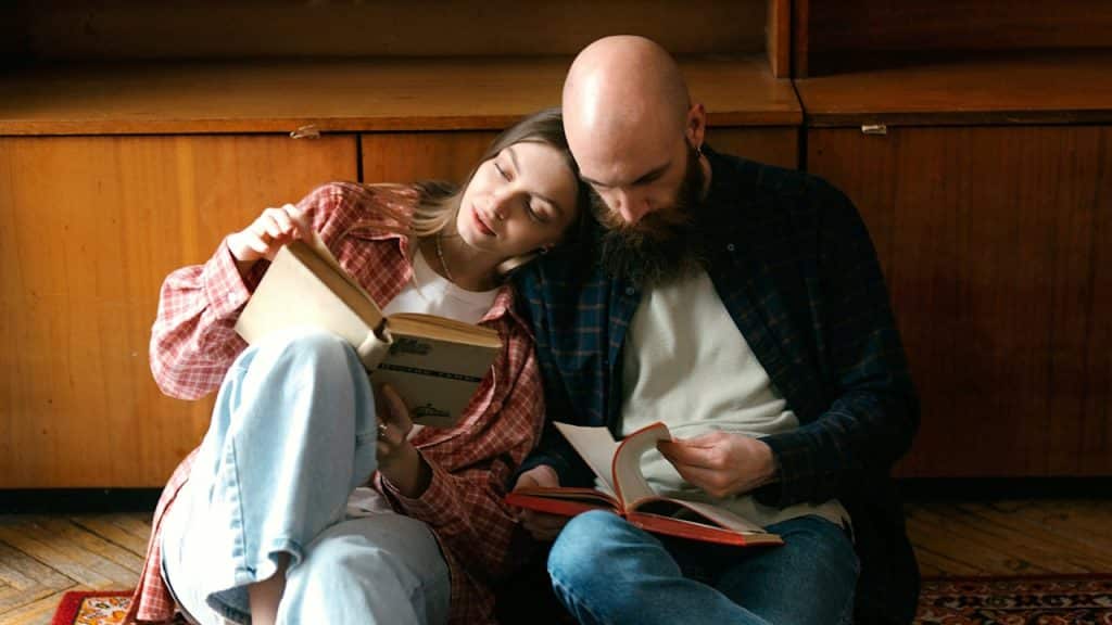 A couple leaning against each other while sitting on the floor and reading books.