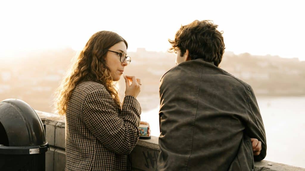 A woman eating ice cream and listening to a man talk with a river in the background.