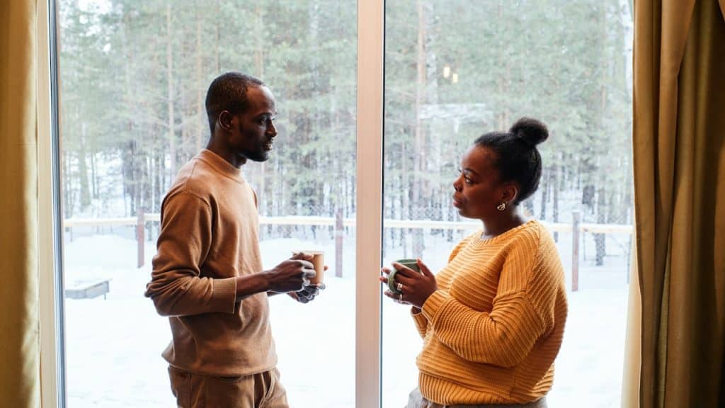A couple having coffee and talking by a window with snowy trees outside.