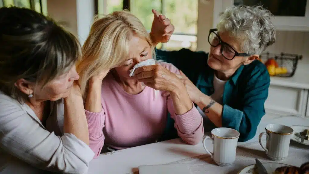 A woman crying at a table while two friends comfort her.