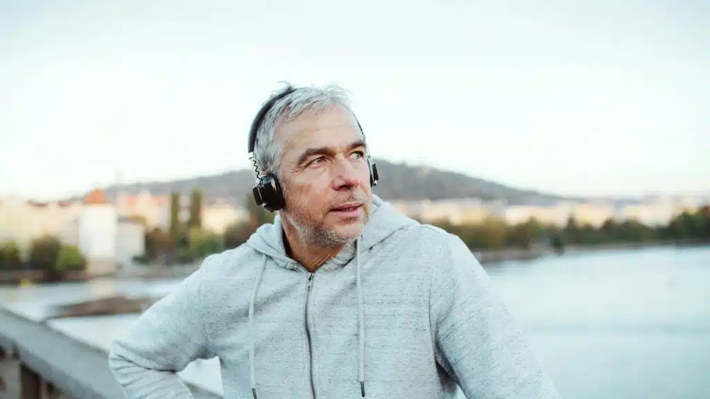 A man wearing headphones and a gray hoodie, standing outdoors by the water.