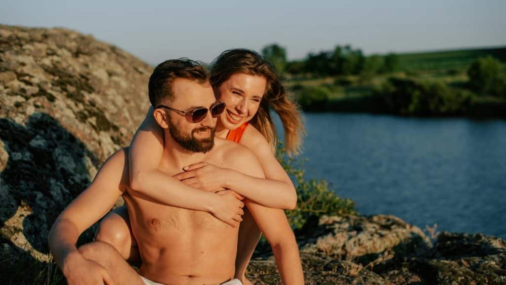 A woman hugging a man from behind while sitting by a lake.