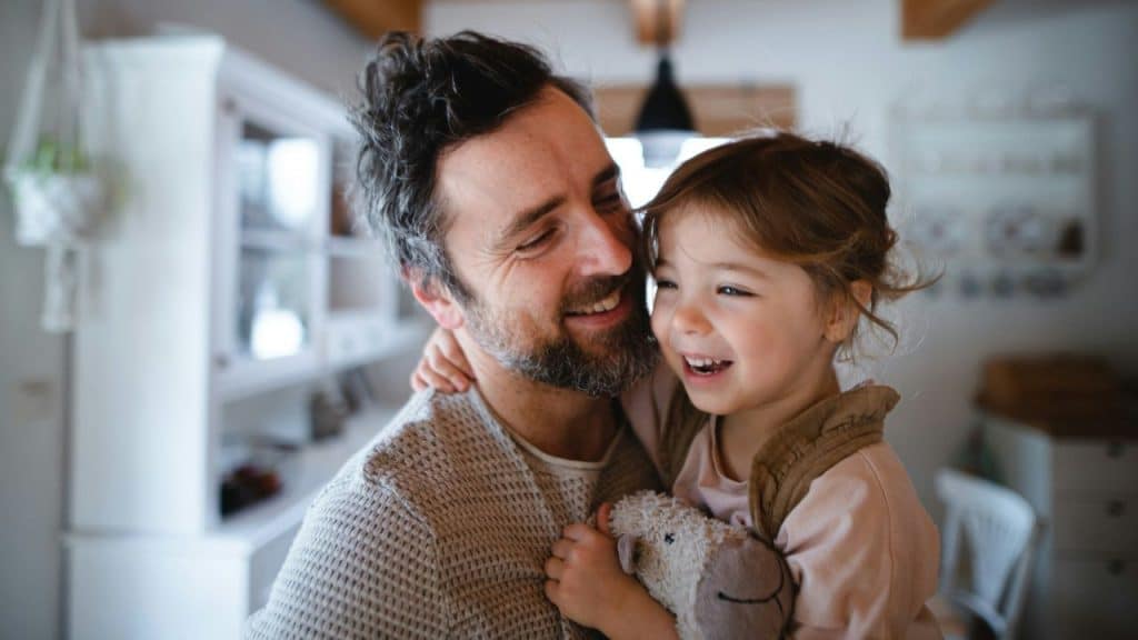 Smiling bearded father hugging his young daughter who is holding a stuffed animal.