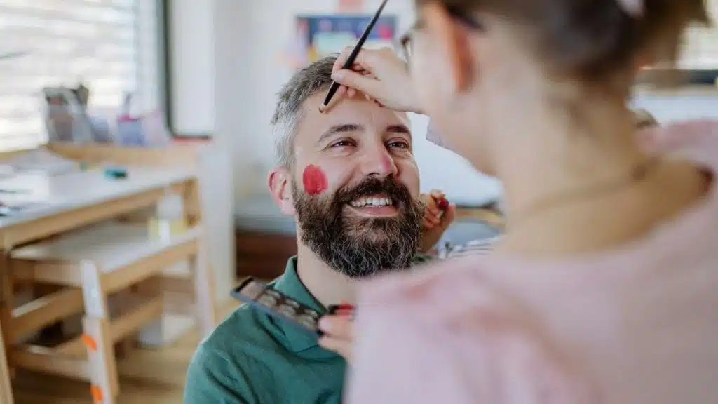 Smiling bearded man getting red face paint applied with a small brush.