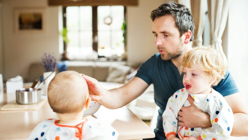 Father feeding one baby at a table while holding another baby with a messy face.