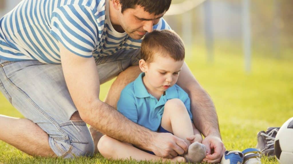 Father in striped shirt kneeling on grass helping a young boy put on a sock.