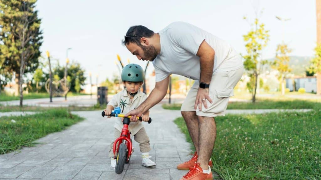 Father bending down to help a small boy in a helmet learn to ride a red balance bike.