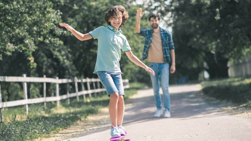 Smiling boy balancing on a skateboard with his father watching and waving behind him.