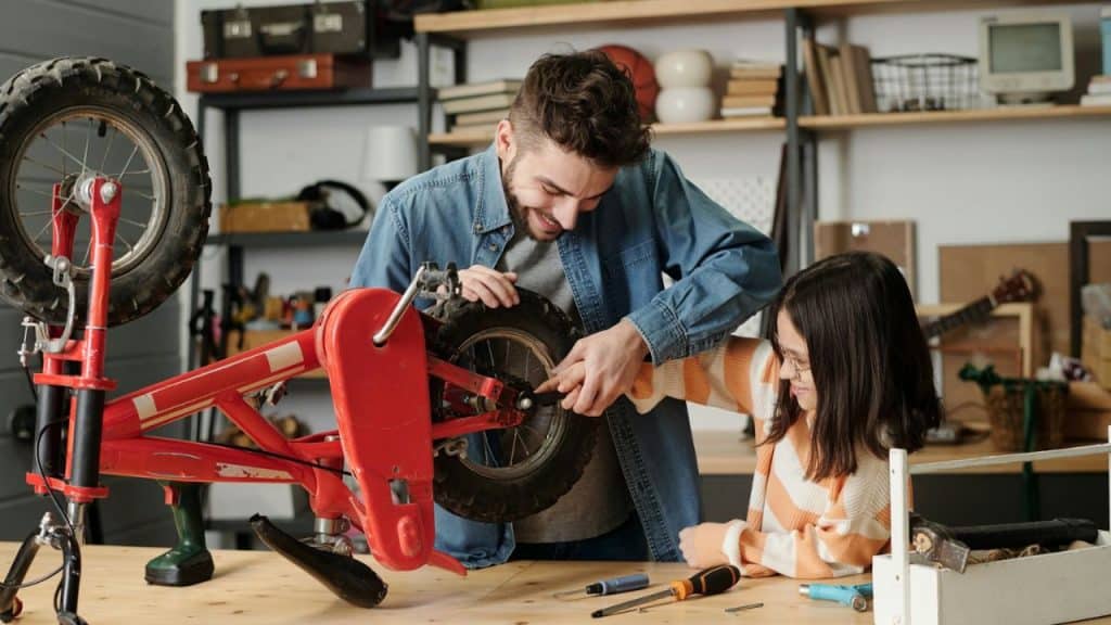 Smiling father and daughter repairing a red bicycle wheel on a workbench in a workshop.