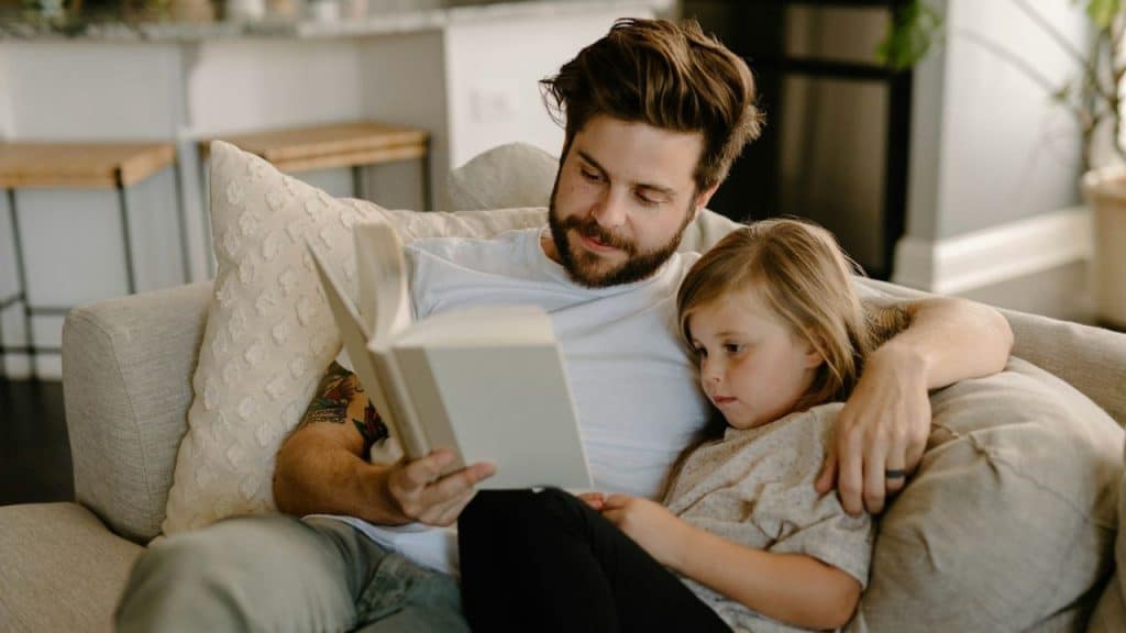 Bearded man with a tattoo reading a book to a young girl on a sofa.