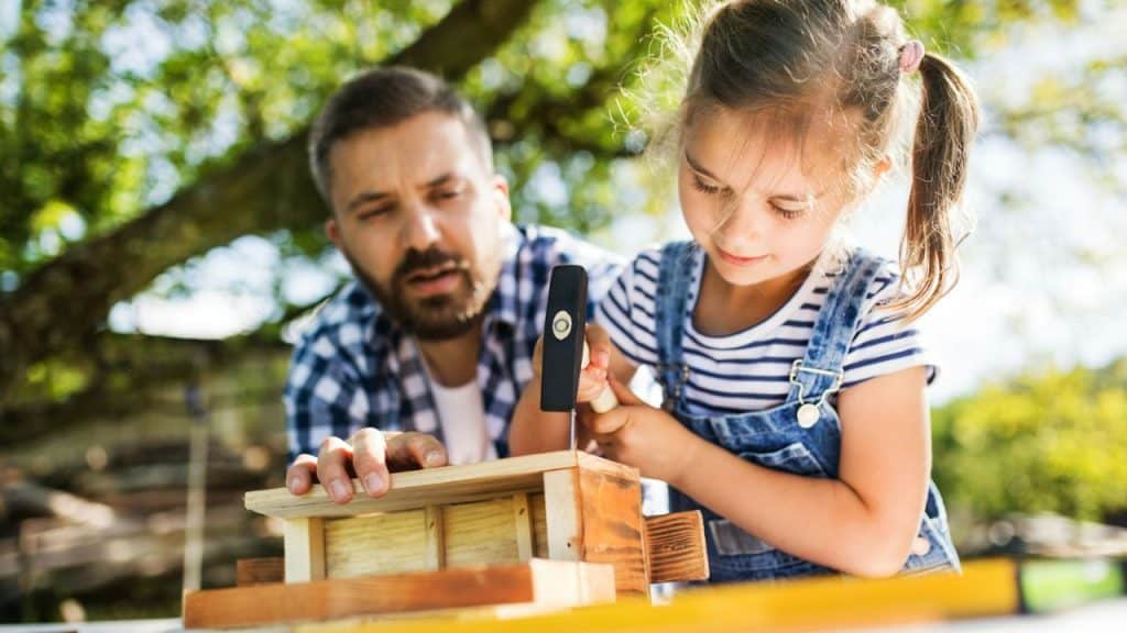 Father in a plaid shirt helping a young girl hammer a nail into a wooden birdhouse outdoors.