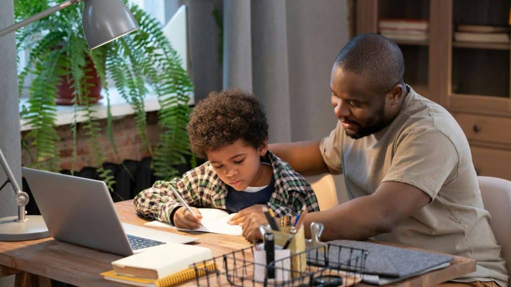 Father with his arm around a young boy who is writing in a notebook at a desk.