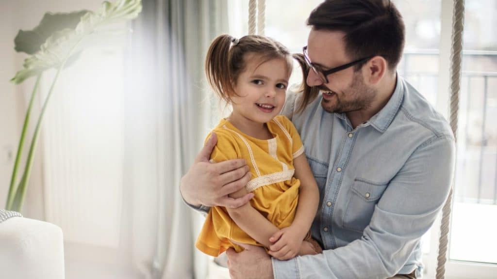 Smiling man in glasses hugging a young girl with pigtails while sitting on a swing indoors.