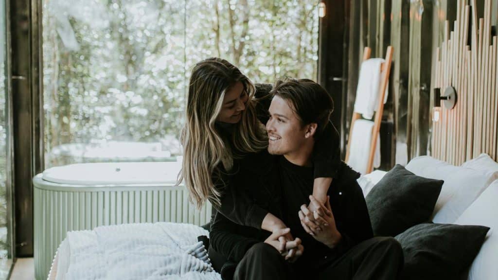 A couple sitting together on a bed smiling at each other.