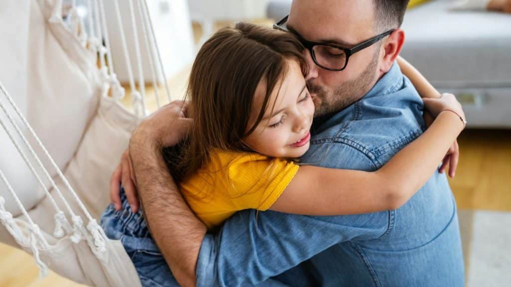 Man in a denim shirt and glasses hugging a young girl who is smiling with closed eyes.