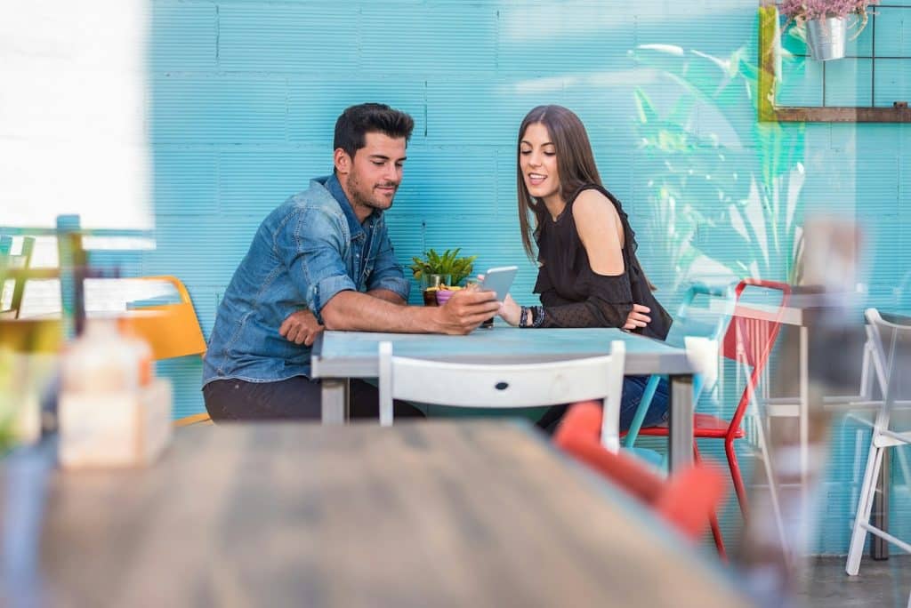 The man is showing his phone to the woman while they’re sitting at a restaurant.