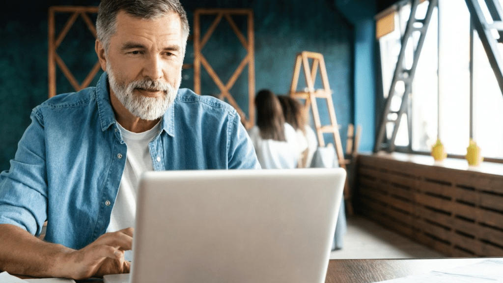 A middle-aged man with a white beard works on a laptop in a modern, teal-walled office.
