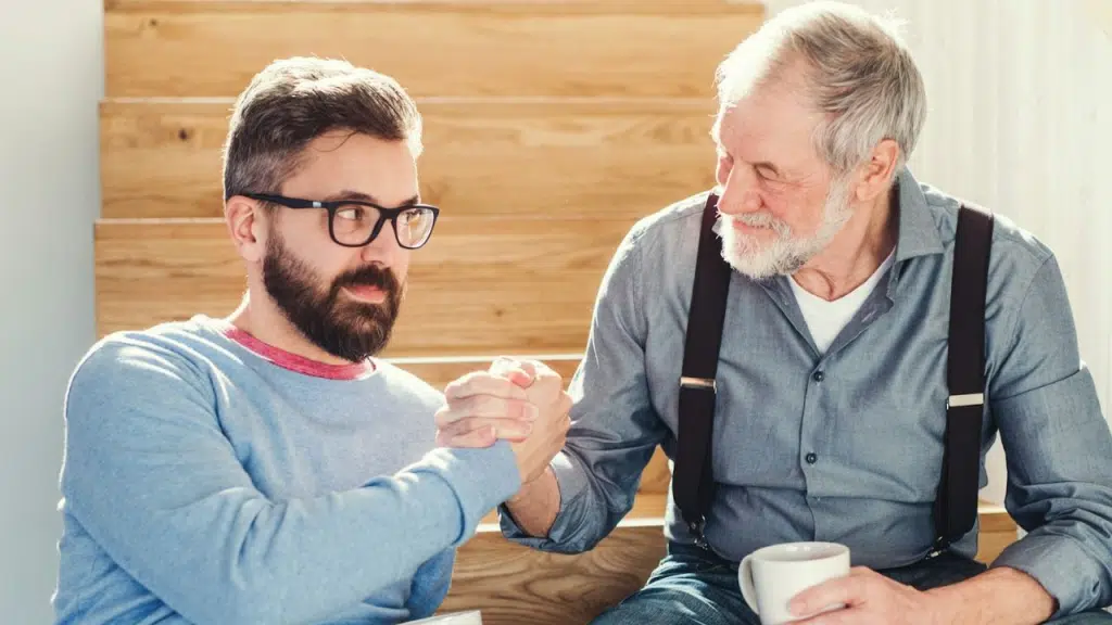 Two men, one older and one younger, are clasping hands while sitting on a wooden staircase.