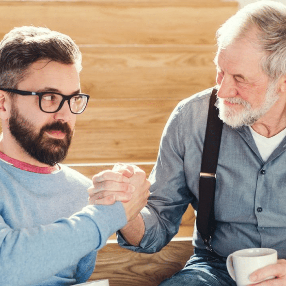 Two men, one older and one younger, are clasping hands while sitting on a wooden staircase.