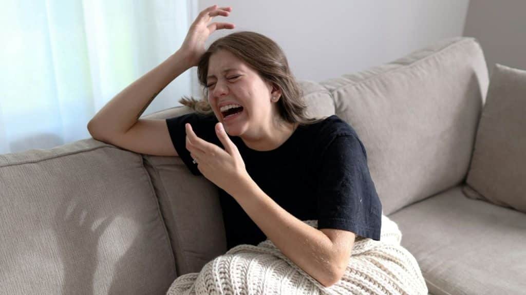 A woman on a beige couch, wearing a black shirt, is visibly upset and crying with her eyes closed and a hand near her face.