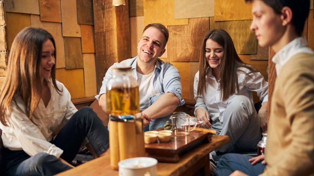 Four adults are sitting around a low wooden table in a warmly lit room with a wood-paneled wall, smiling and laughing together.