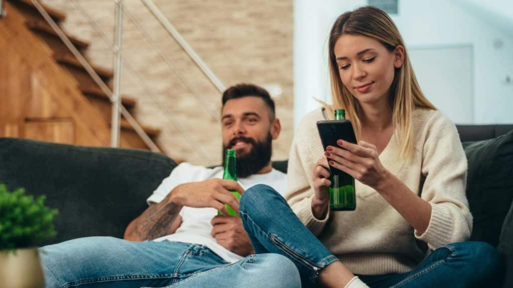 A woman scrolls her phone while holding a green bottle; a smiling man with a beard sits beside her, also holding a green bottle.