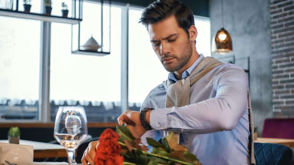 A man in a blue shirt checks his wristwatch at a restaurant table, next to red roses and a wine glass.