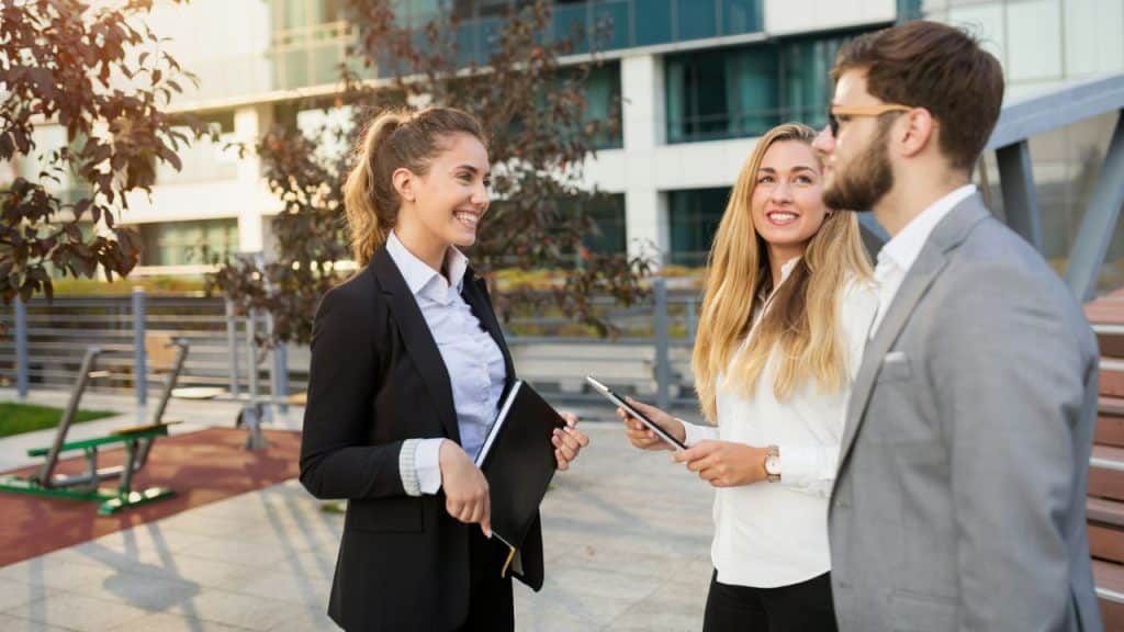 Three professionally dressed individuals are standing outside a modern building, smiling and talking during the day.
