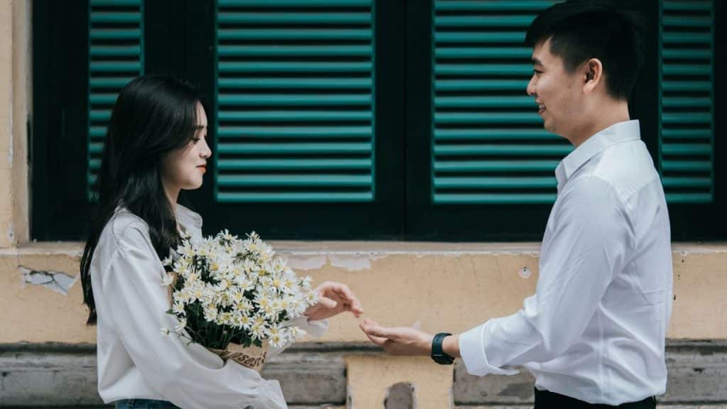 A man and woman in white shirts stand facing each other, exchanging a bouquet of white flowers against a yellow wall.