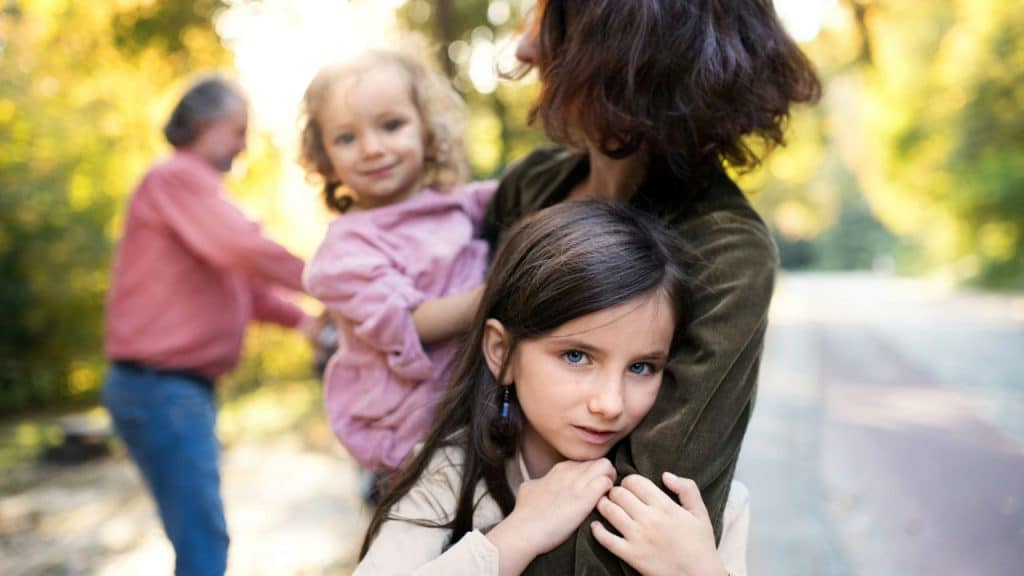 A woman holds two young girls in an outdoor park setting with blurred background and bright sunlight.