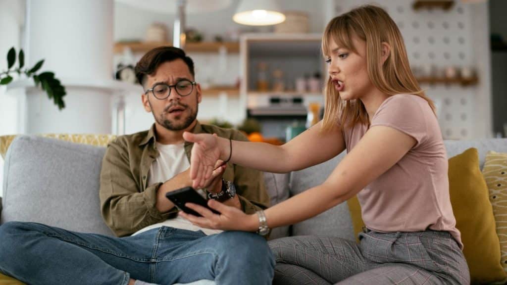 A woman holding a smartphone sits on a couch, gesturing at a man with glasses who looks surprised or confused.