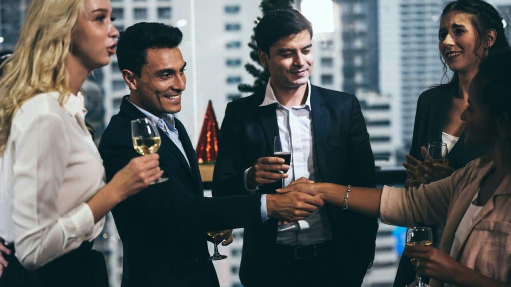 Five professionally dressed people at a nighttime gathering, shaking hands and holding glasses of wine.