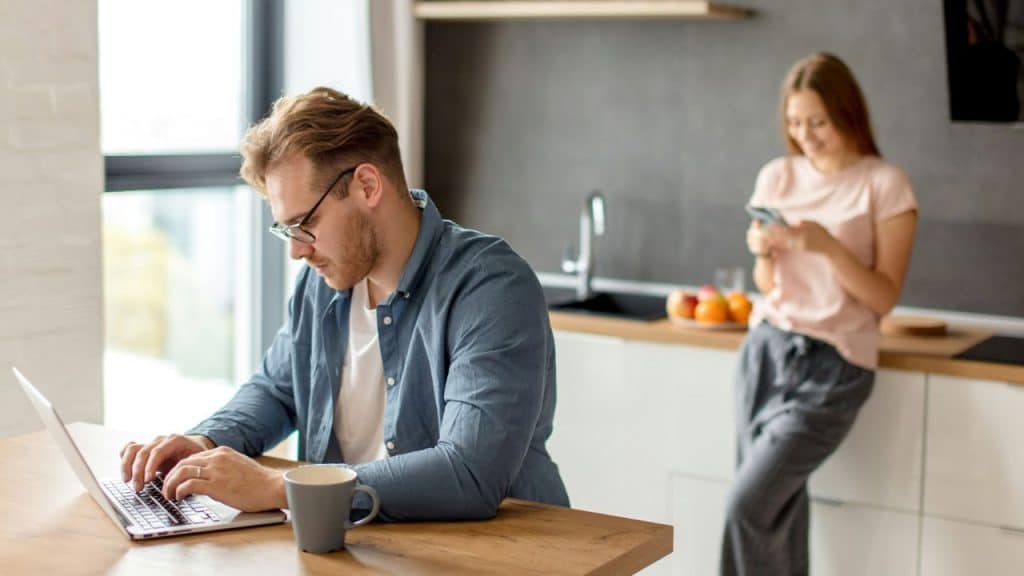 A man wearing glasses works intently on a laptop at a kitchen table while a woman stands in the background looking at her phone.