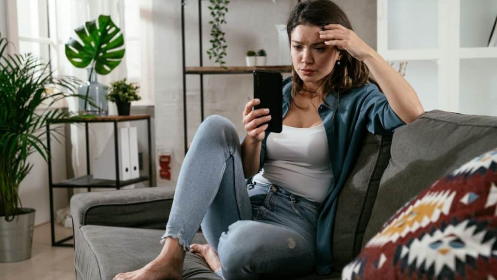 Woman on a sofa looking stressed at her phone with her hand on her forehead.