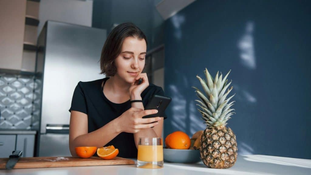 Young woman smiling while looking at her phone at a kitchen counter with fruit.