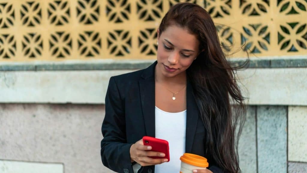 Young woman with long dark hair looking at her phone, holding a coffee cup.