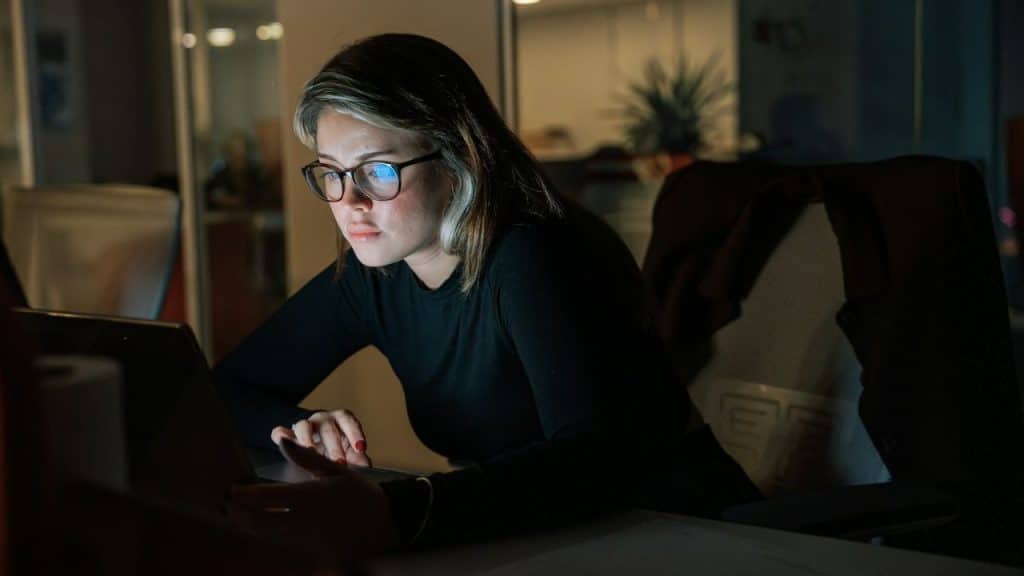 Woman wearing glasses and a black shirt works on a laptop in a dark office.