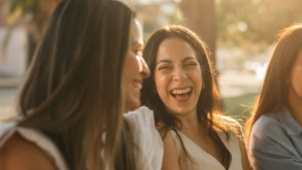Two women outdoors, one in the center laughing brightly with her mouth wide open.