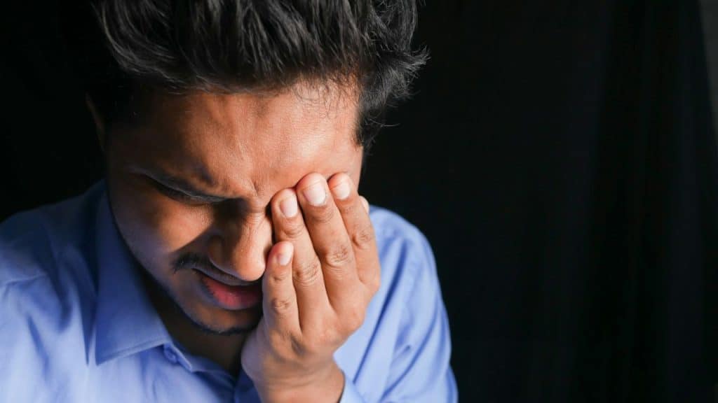 Close-up of a distressed man rubbing his eyes, wearing a blue collared shirt.