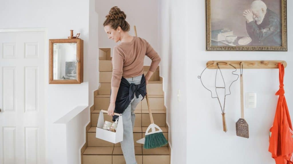 Woman holding a cleaning caddy and a broom, walking up a staircase inside a home.